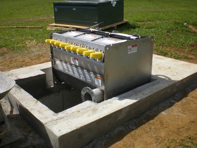 Uma unidade da Chave de Distribuição Subterrânea Vista® está sendo instalada em uma base de concreto em primeiro plano. O interior de metal selado da unidade está exposto, e o gabinete verde que o a cobrirá está em um pallet de madeira na grama em segundo plano. A unidade tem nove buchas salientes cobertas por tampas amarelas. Sua base repousa sobre uma parte escavada da plataforma de concreto e, abaixo dela, há um outro espaço escavado de profundidade desconhecida. 